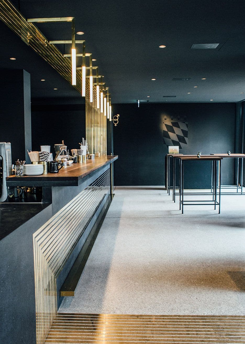 Modern dark bar interior with a wooden counter, gold accents, and hanging tube lights.