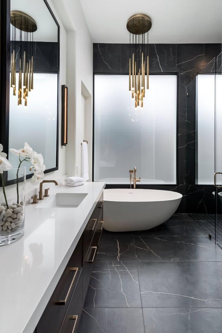 Modern bathroom featuring black marble tiles, white freestanding tub, and elegant gold chandelier lighting.