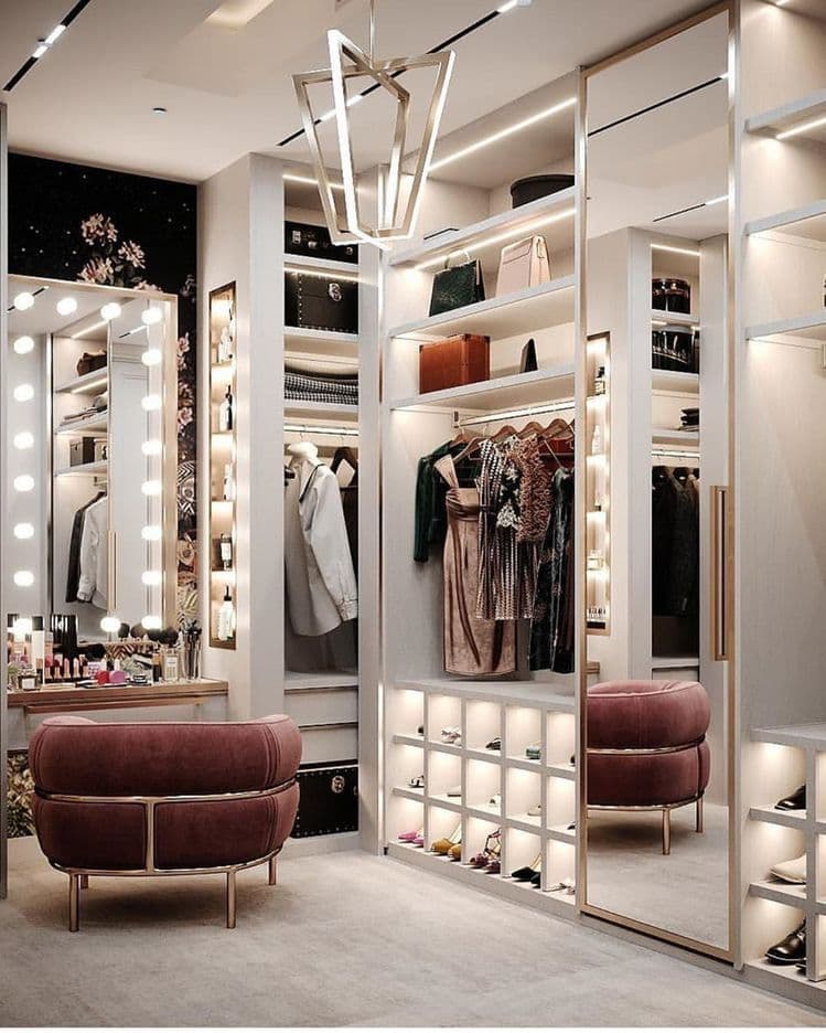 Luxurious walk-in closet with white shelving, a lit vanity, and a plush mauve armchair.