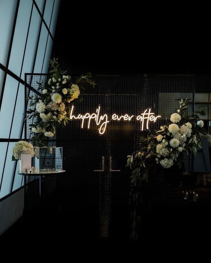 Neon 'happily ever after' sign on black mesh backdrop adorned with white floral arrangements.