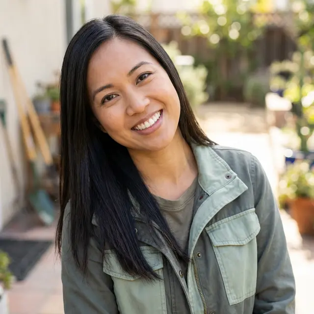 Smiling woman with long dark hair wearing a green jacket in a sunny garden.