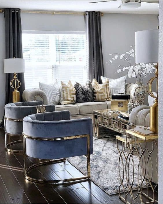 Modern living room with blue velvet armchairs, gold accents, grey sofa, and mirrored coffee table.