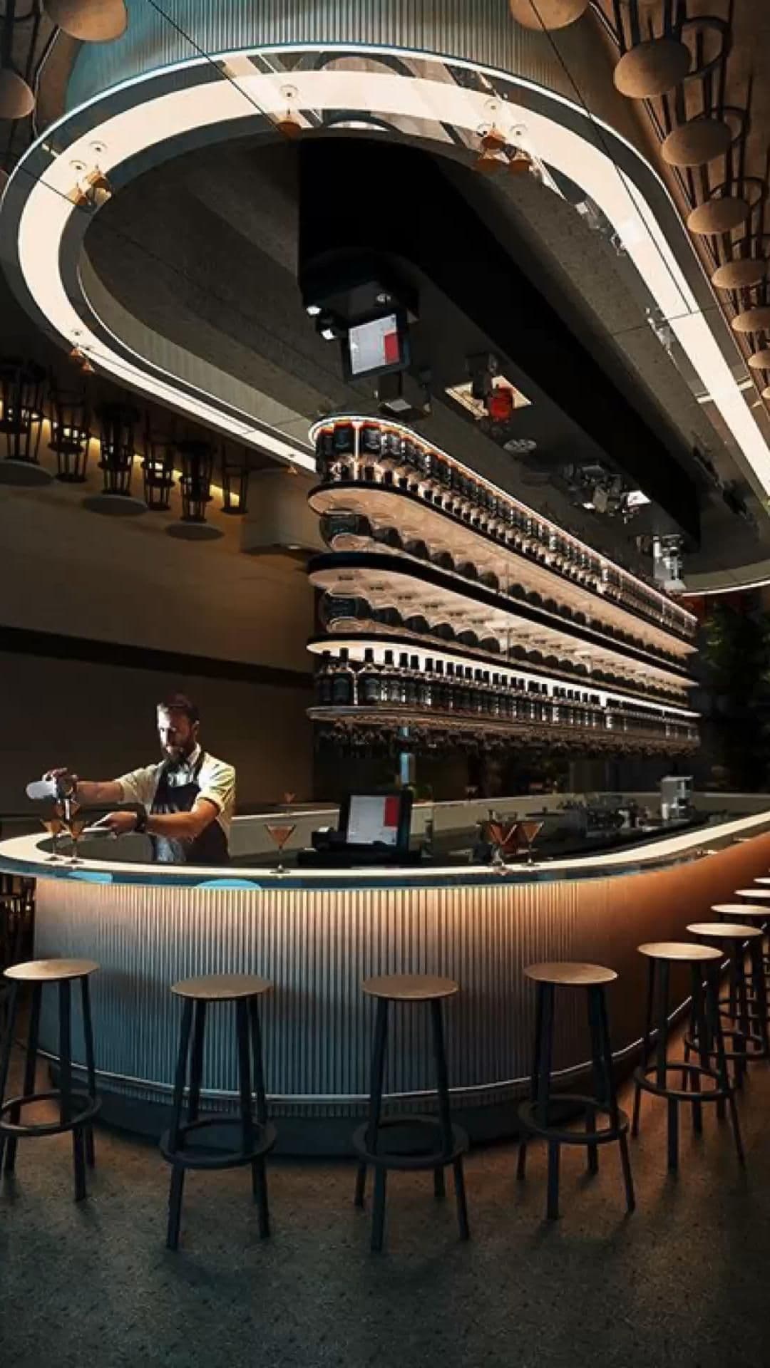 Bartender mixing drinks at a modern curved bar with tiered bottle shelves and stools.