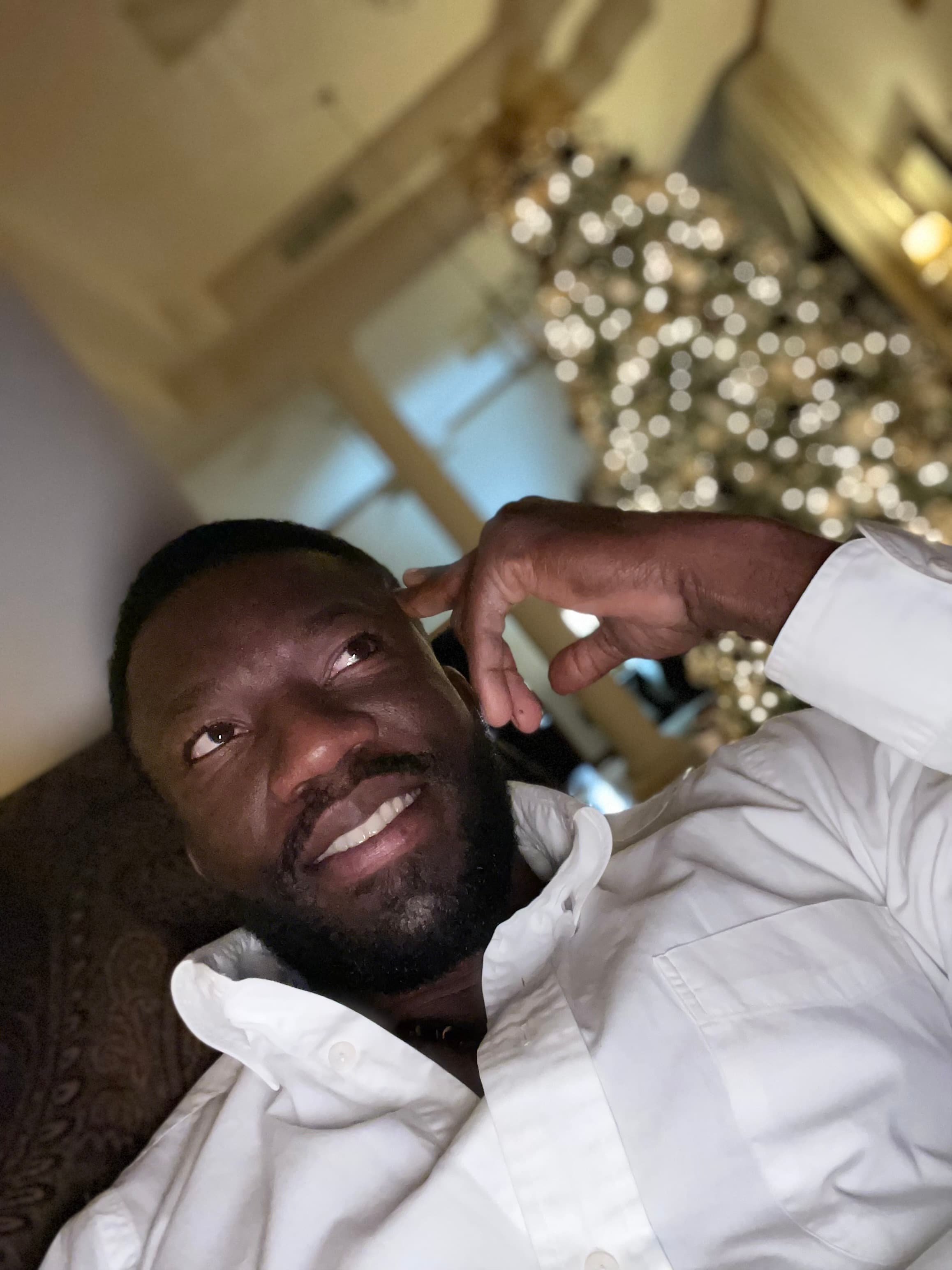 Smiling bearded Black man in a white shirt looking up near a blurred Christmas tree.