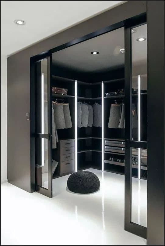Sleek black walk-in closet with vertical LED light strips, glass doors, and a round ottoman.